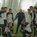 A soccer coach gives tactical advice to a youth team indoors on artificial turf.
